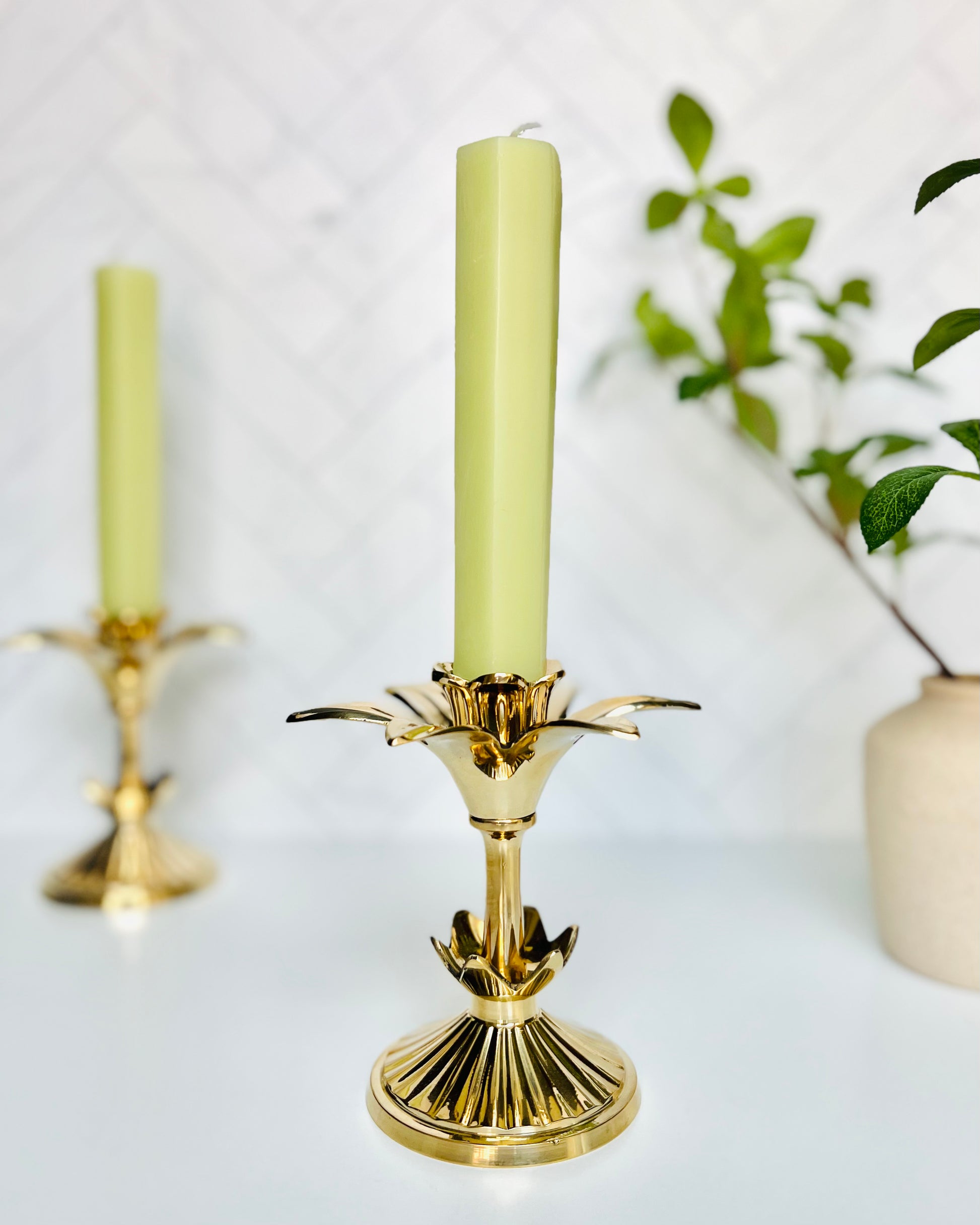 A melon taper candle in a golden flower candlestick with another one in the background along with a vase filled with greenery