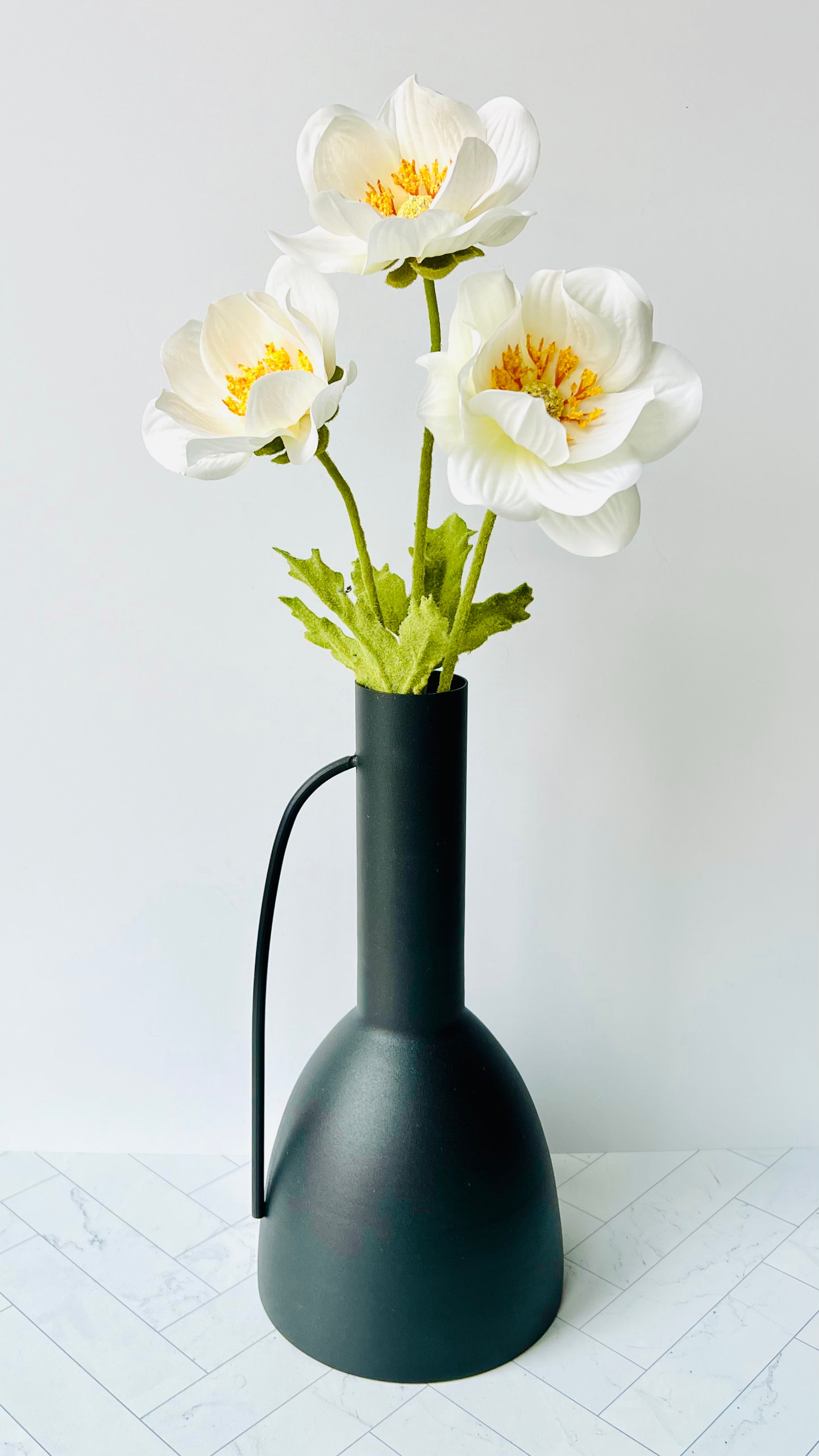 The Sleek Black Vase on a white tiled surface filled with white and yellow flowers