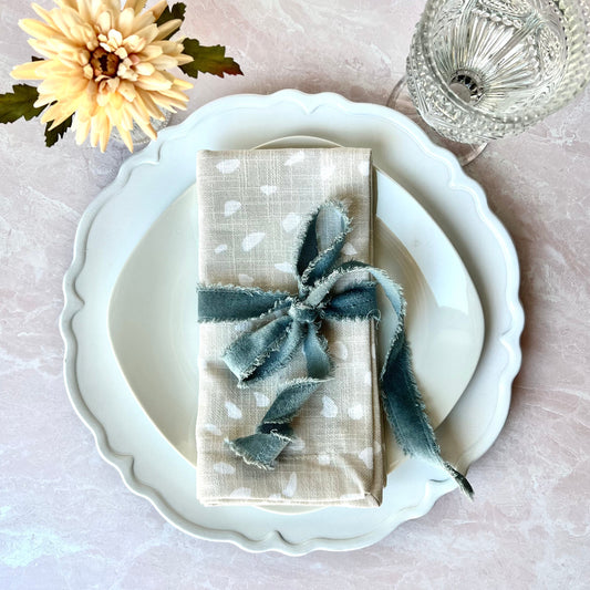 A Fawn Print Napkin shown overhead on a white plate wrapped in a blue velvet ribbon