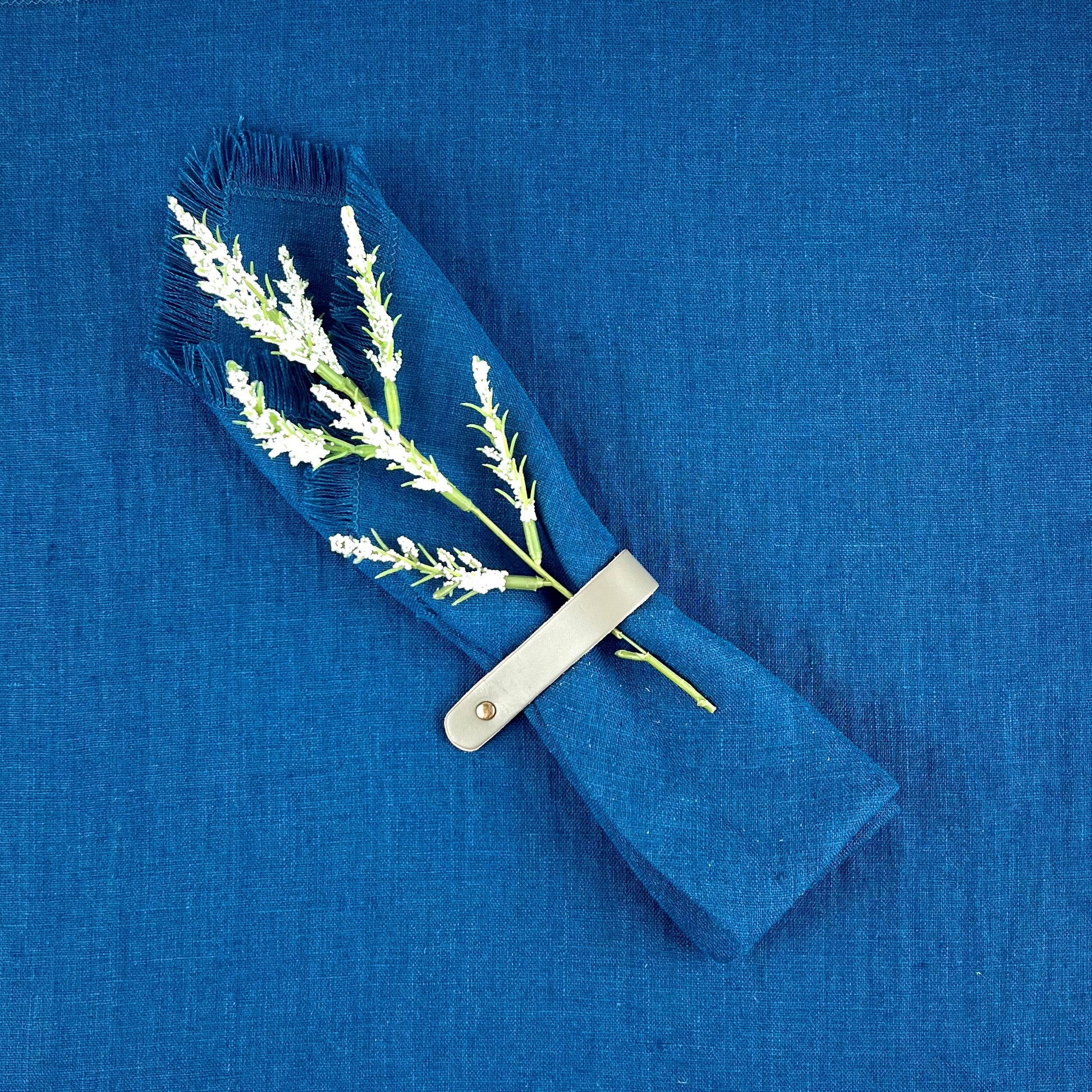 Deep Blue Linen Napkins with one folded on top with a plant sprig tucked in to a gray leather napkin ring